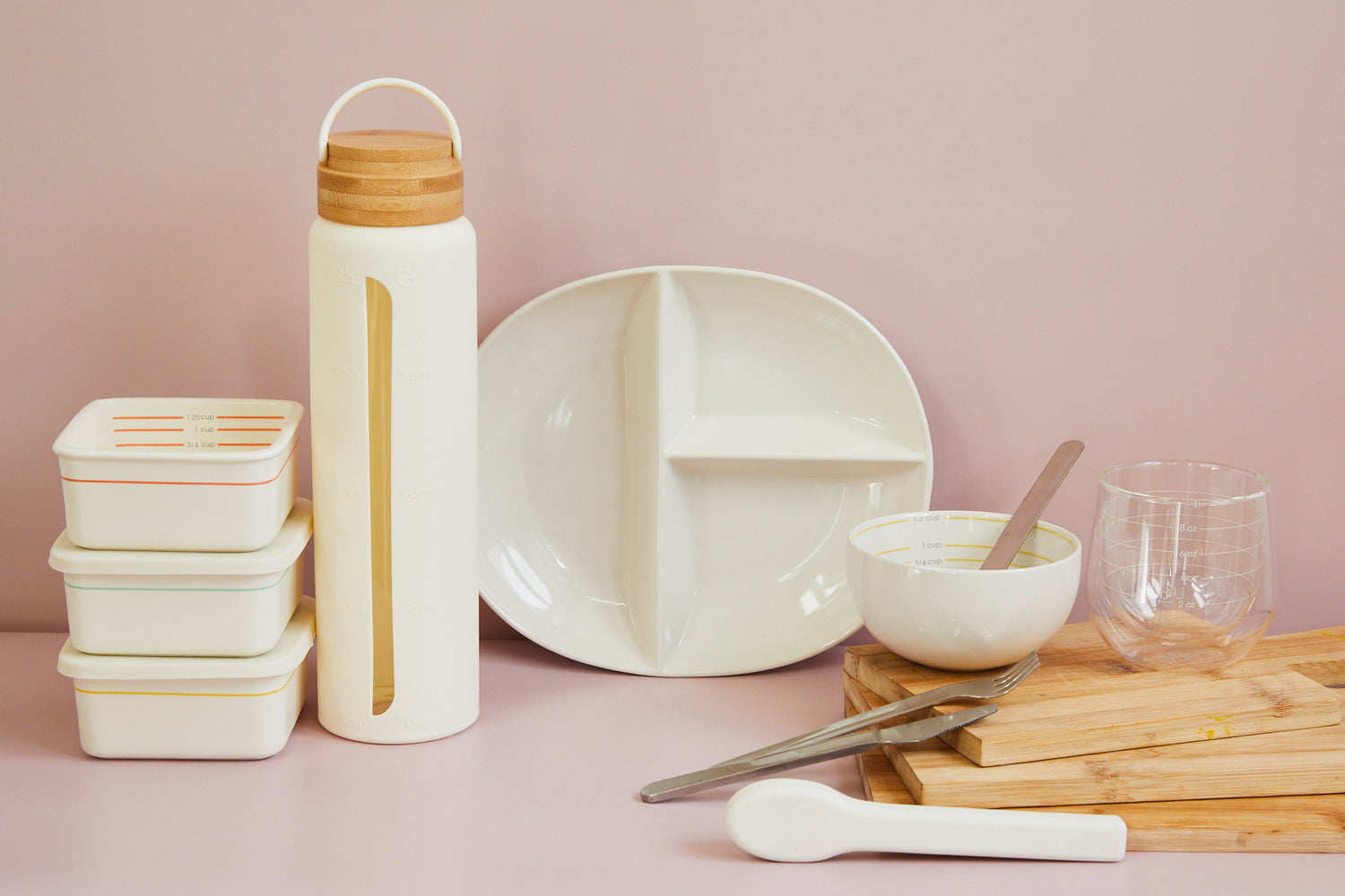 product photography with portion control tools: 3 containers stacked, 1 water bottle, 1 portion plate, 1 bowl with a spoon inside, fork and knife laying over 3 stacked cutting boards and a glass on top of the boards
